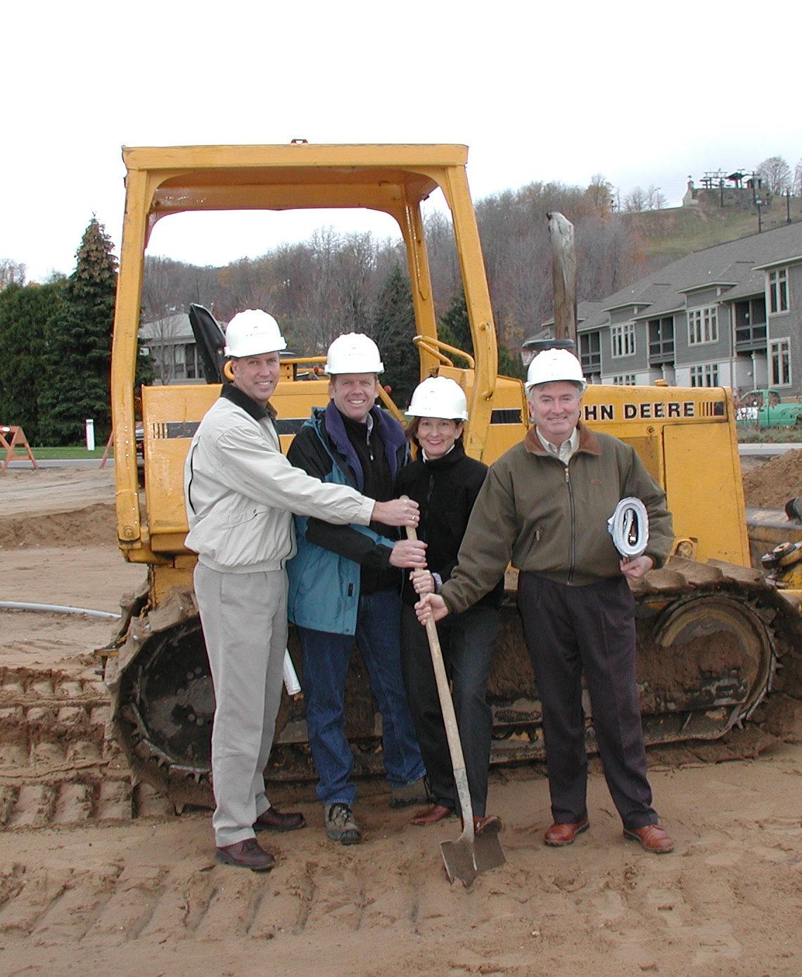Four people in hard hats stand with a shovel in front of a yellow bulldozer.