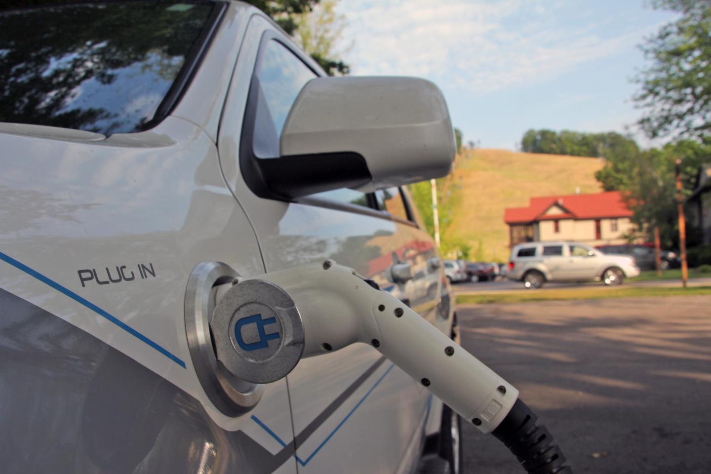 Electric car charging outdoors near houses and trees.