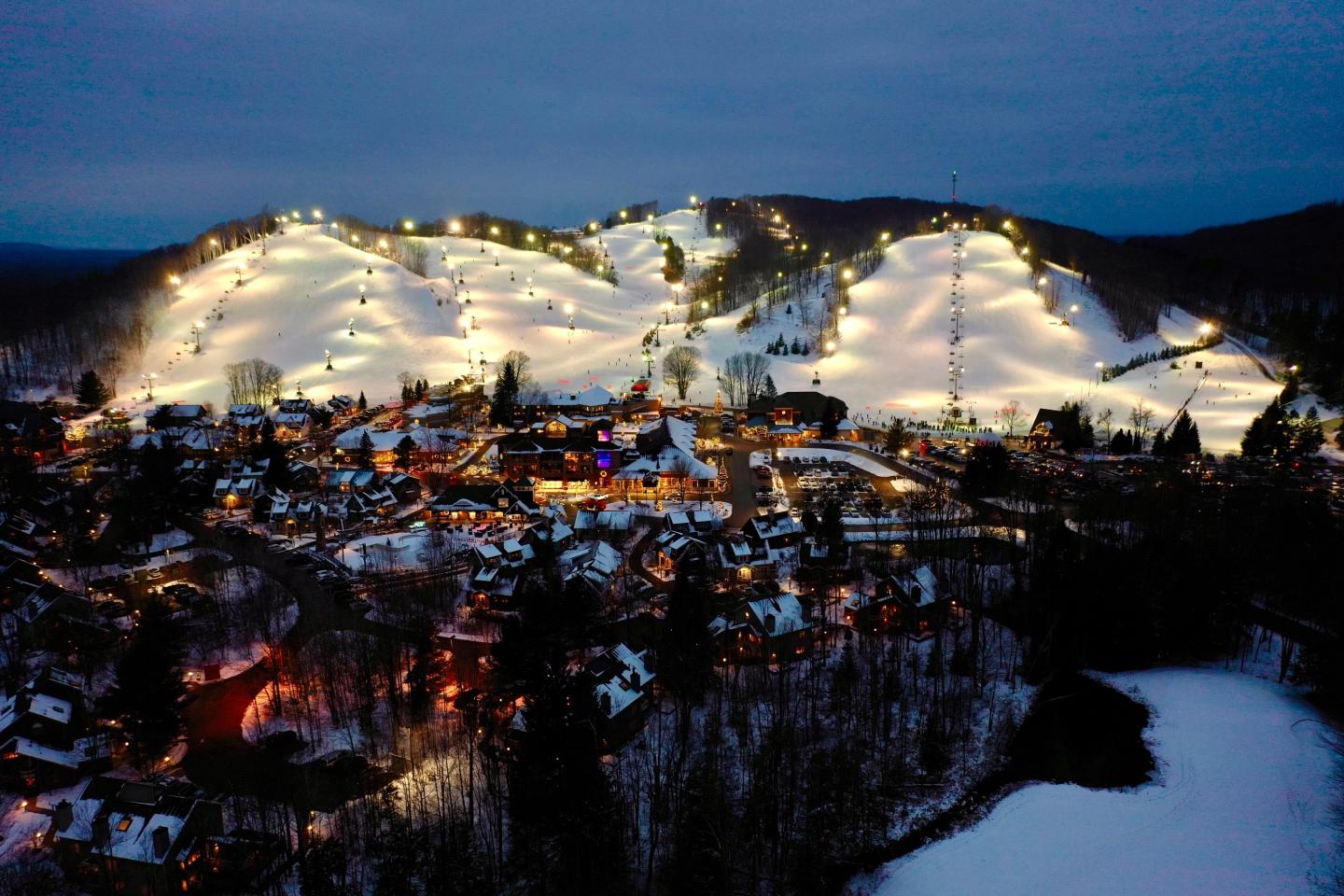 Snowy ski slopes lit at night, overlooking a small town.