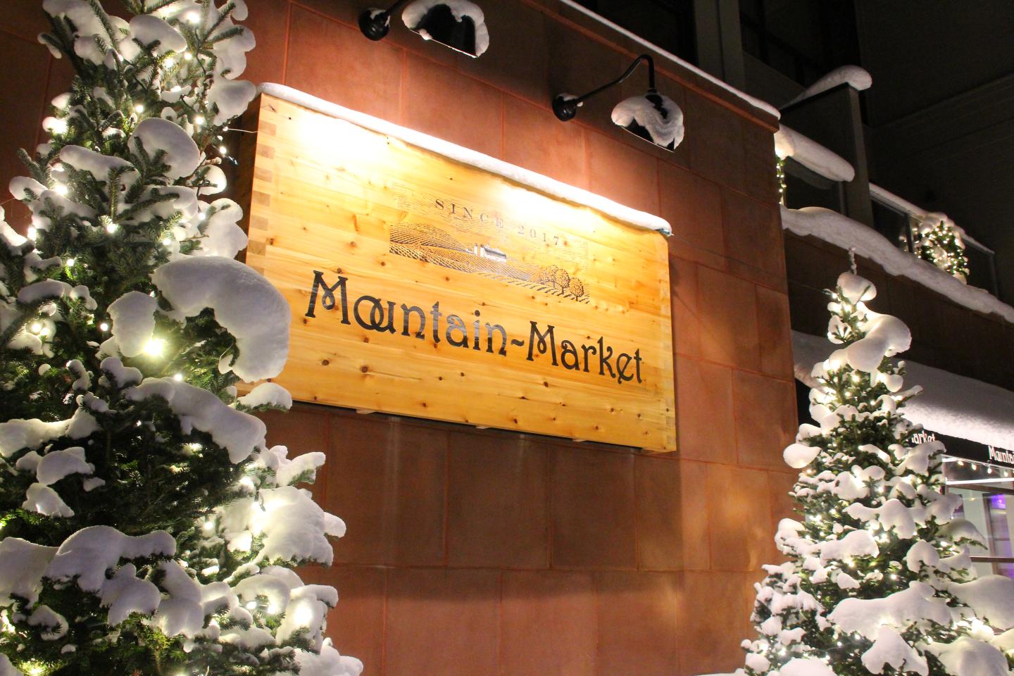 Snow-covered trees with lights beside wooden market sign at night.