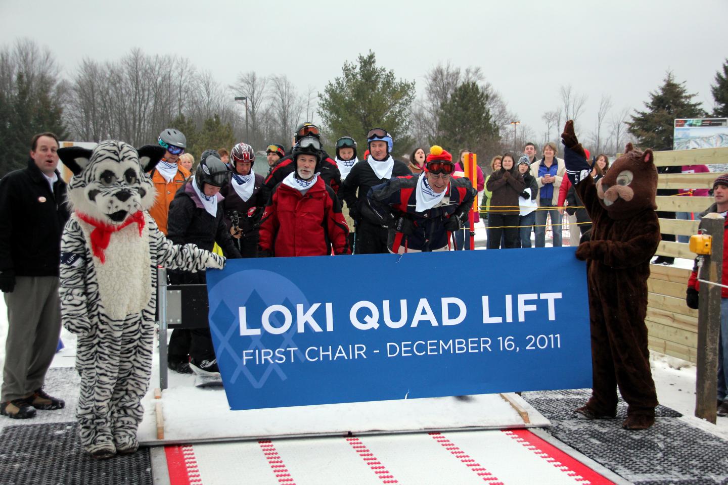 A group of people in winter gear hold a banner near a ski lift, flanked by costumed characters.