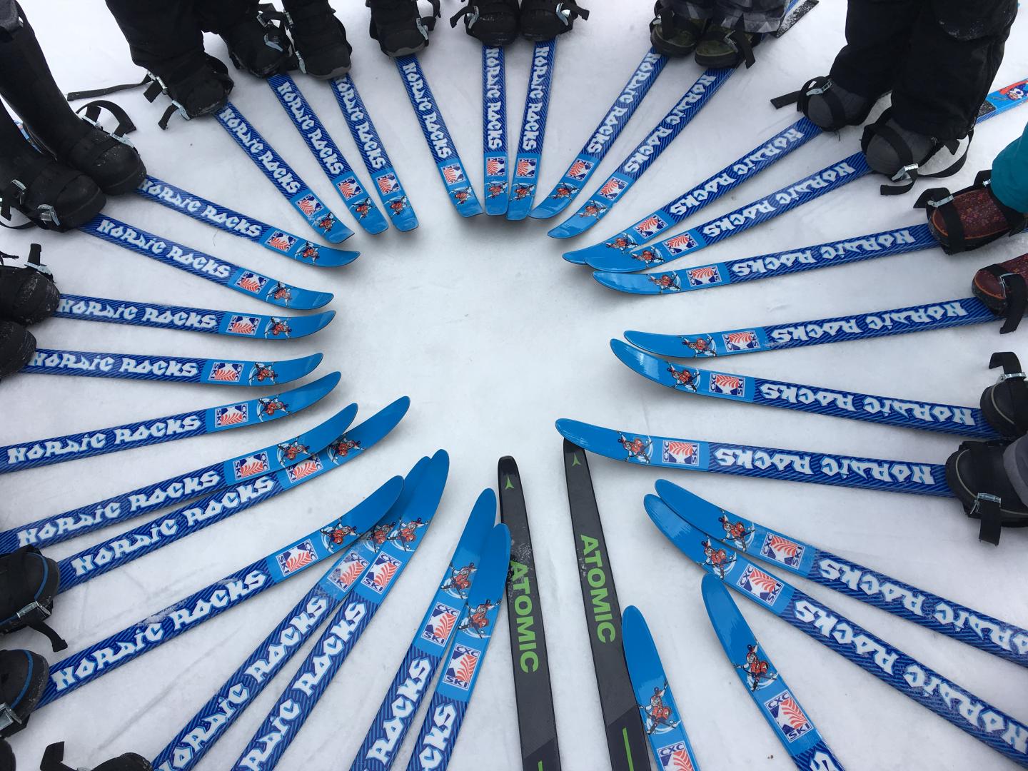 Blue skis arranged in a circle on snow, with a variety of ski boots.