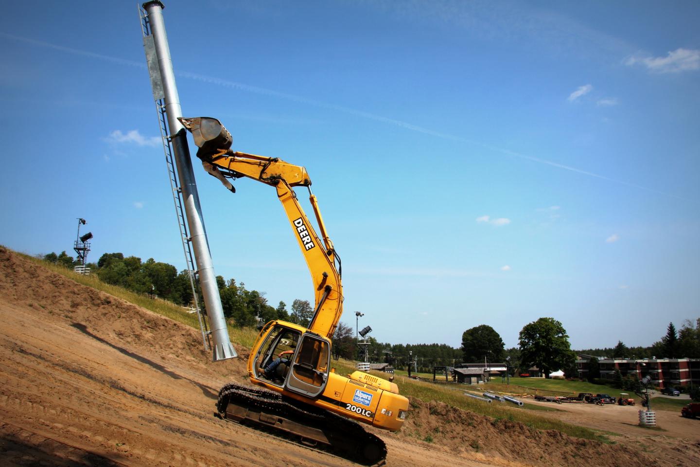 Yellow excavator on a hill, lifting a large metal pole under clear blue sky.