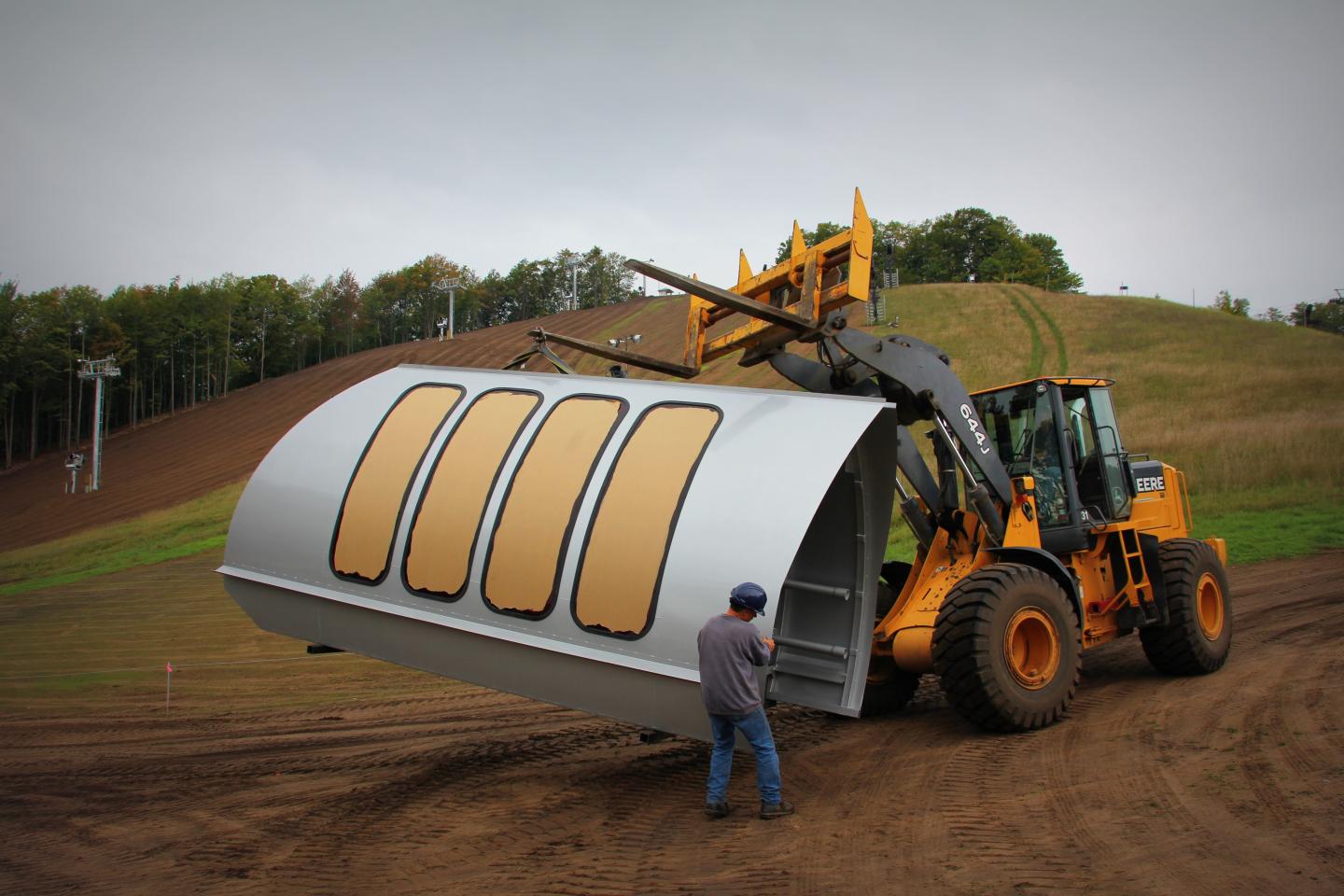 A forklift carries a large, curved metal structure on a dirt hill.