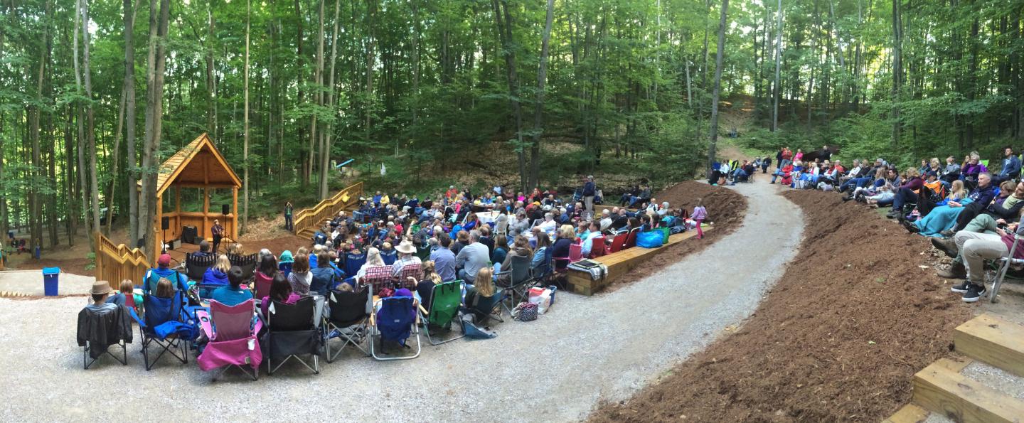 Outdoor amphitheater with audience seated, surrounded by trees.