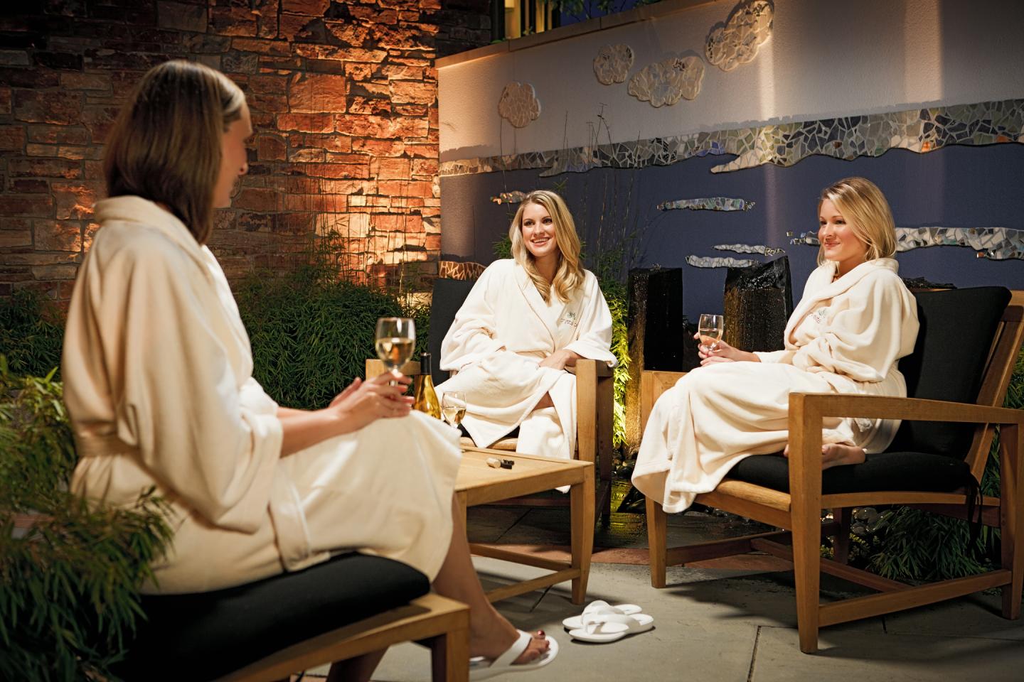 Three women in robes relax in a cozy lounge, holding glasses and smiling.