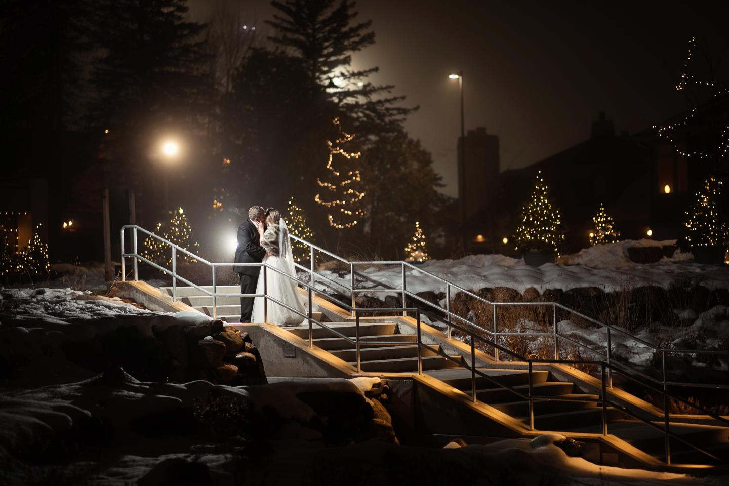 Bride and groom embrace on a snowy, lit-up bridge at night.