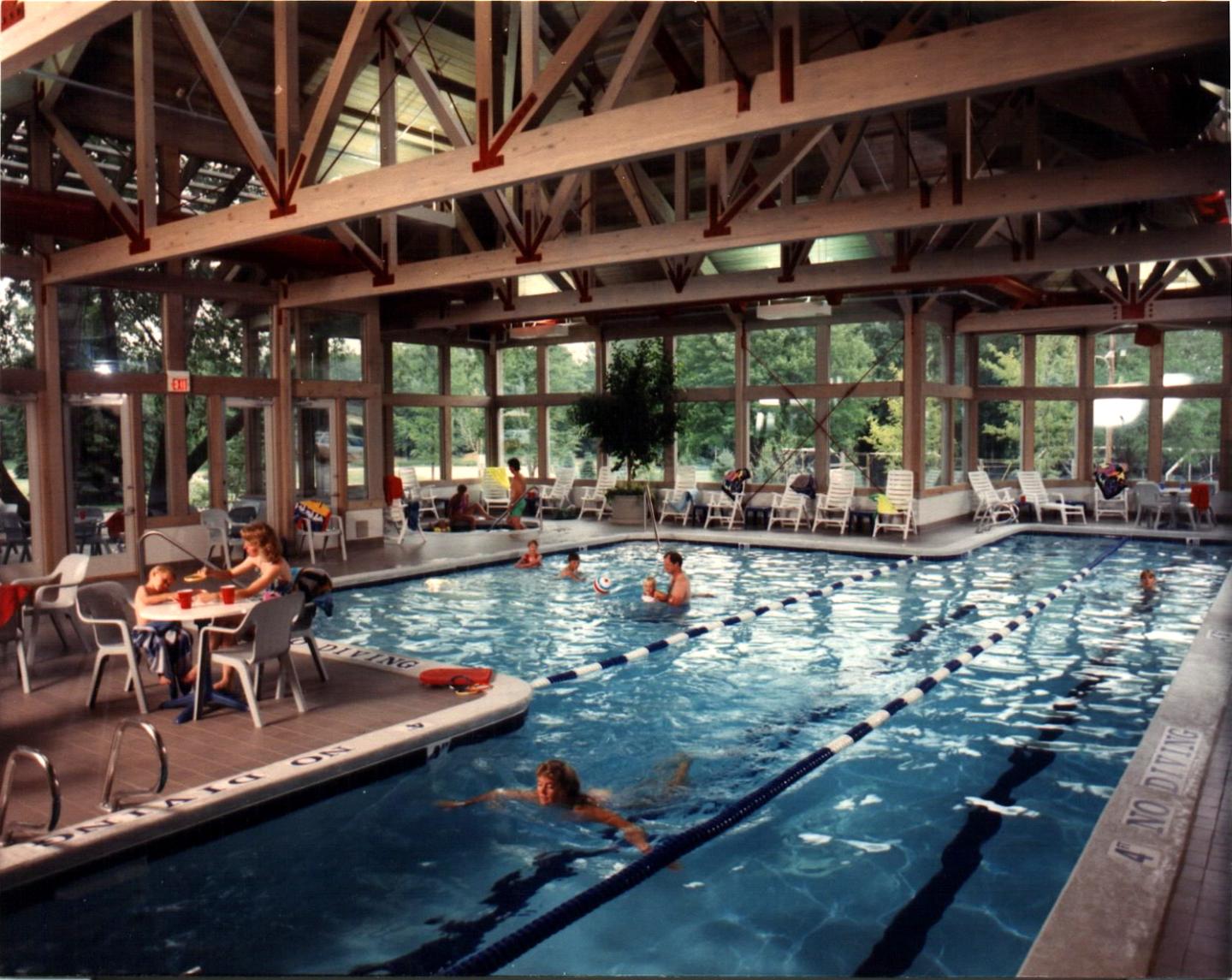 Indoor pool with swimmers, tables, and large windows with a garden view.