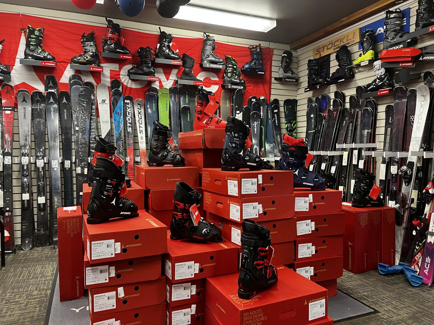 Ski boots on red boxes, surrounded by skis in a store.