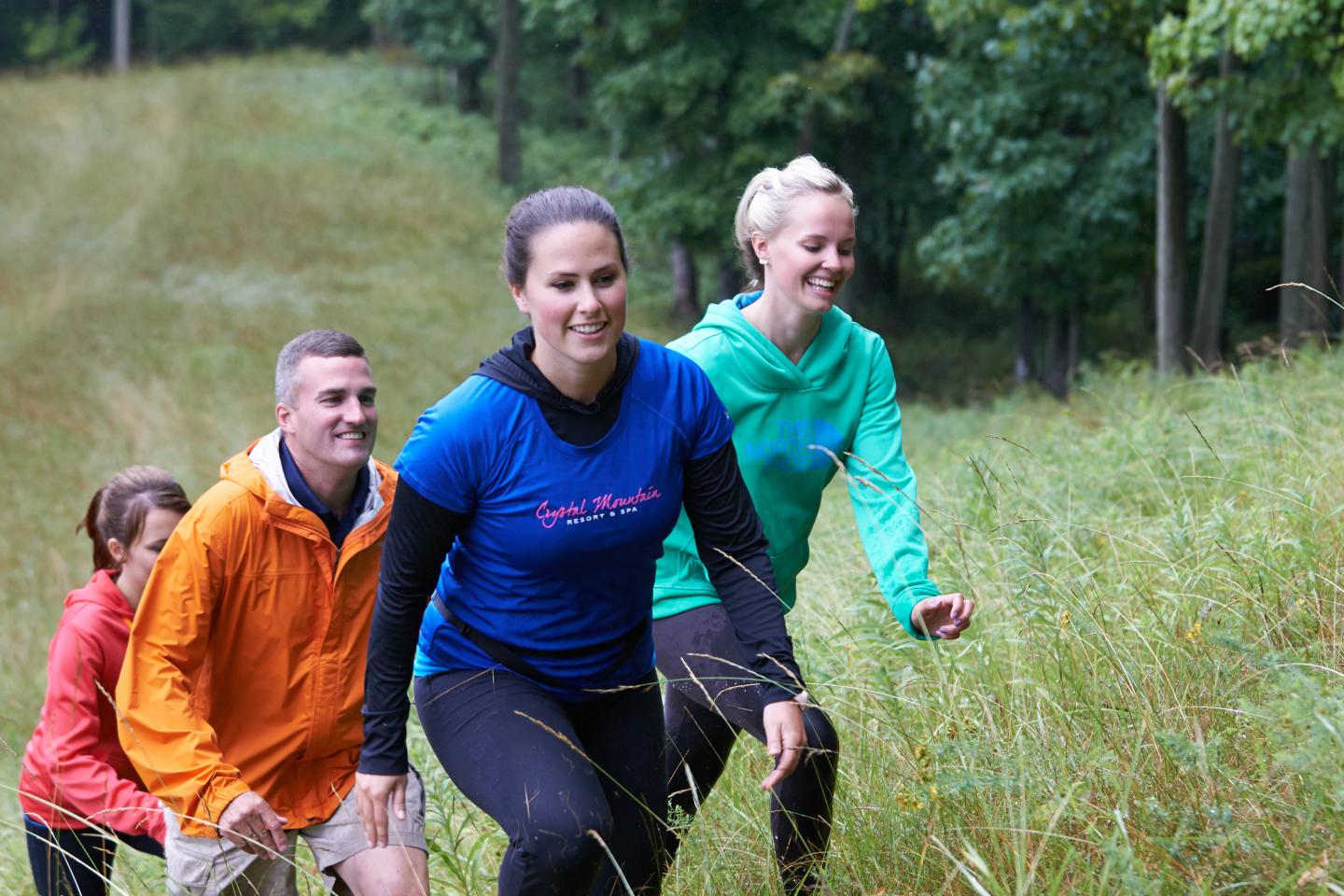 Group hiking uphill through grassy field, smiling and dressed in colorful jackets.