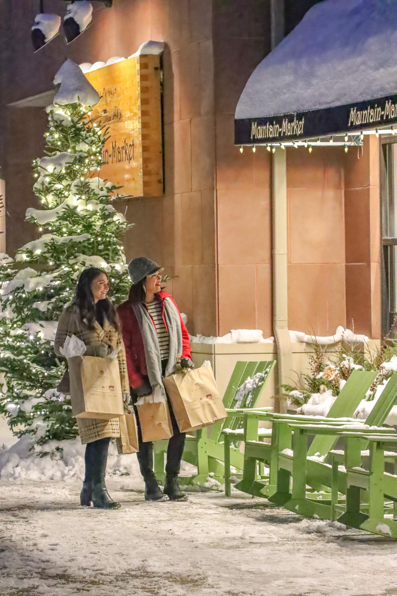 Two women with shopping bags smile outside a snowy store with a lit Christmas tree.