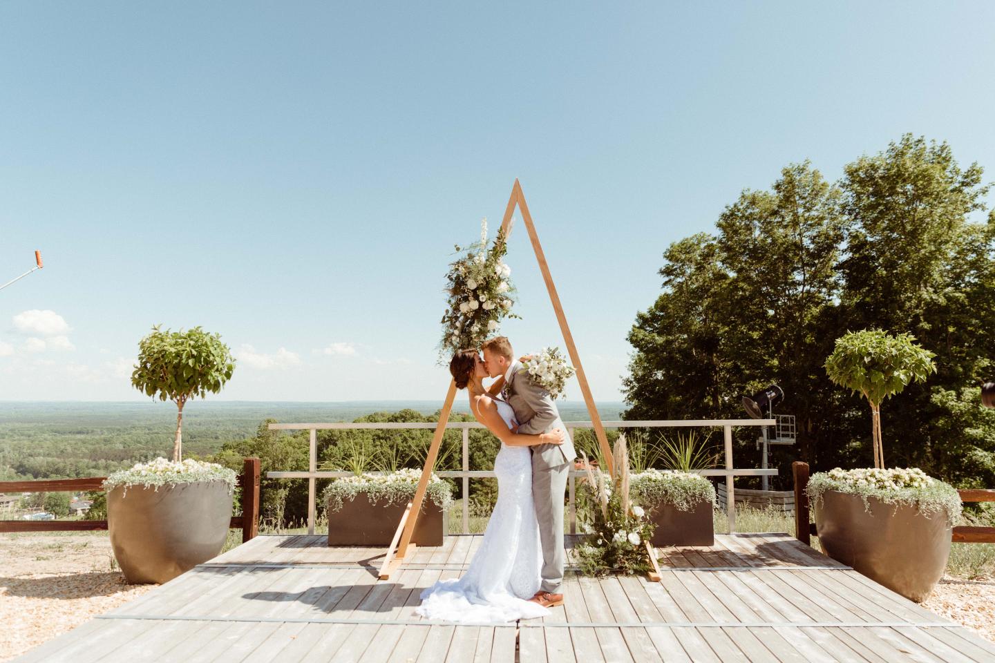 Bride and groom kissing under arch, outdoors with clear sky and trees.