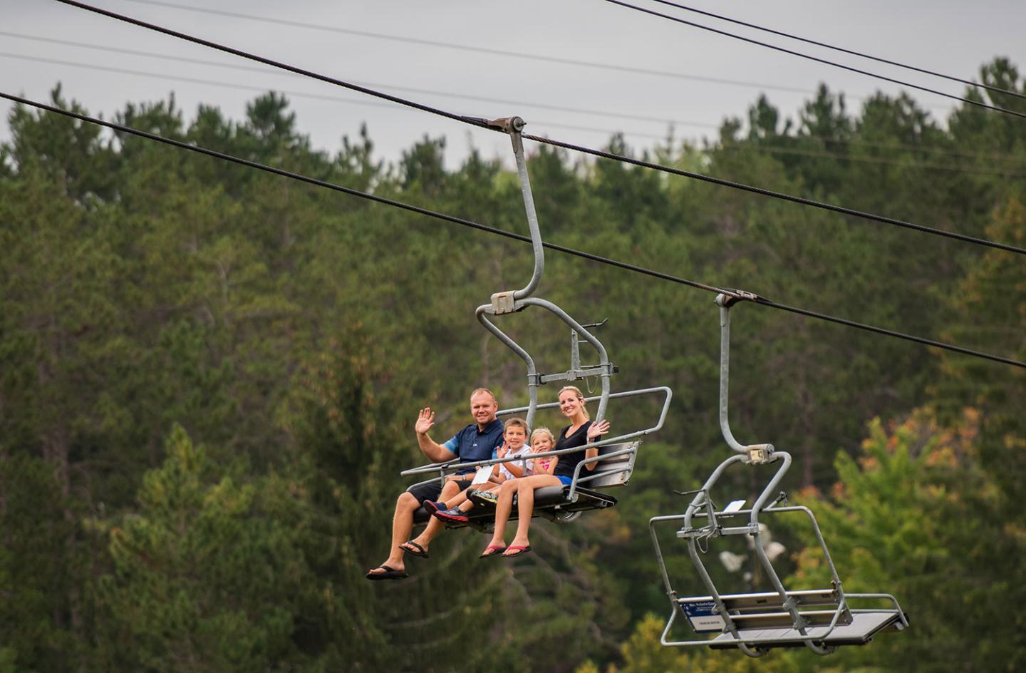 Family on a ski lift, waving, with forest background.