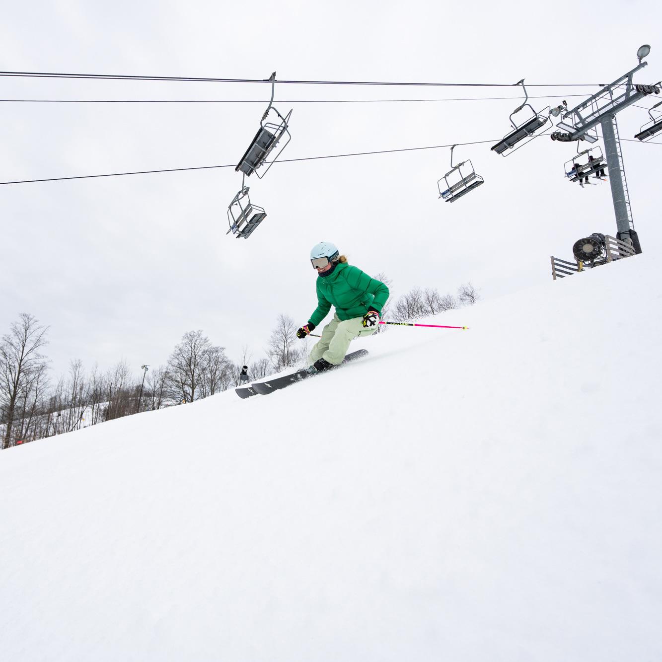 Skier in green jacket descending snowy slope beneath ski lifts.