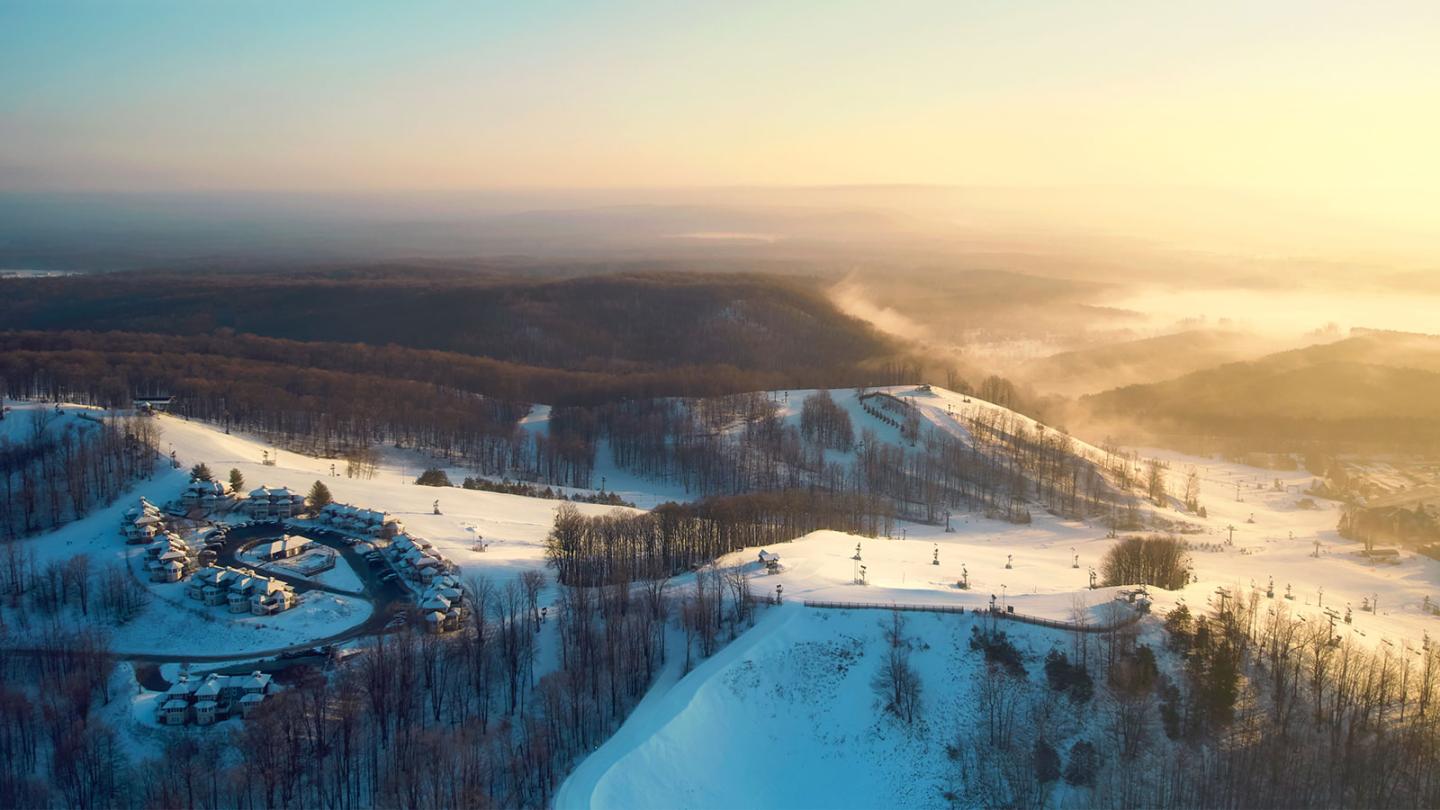 Snowy landscape with a ski resort at sunrise, surrounded by forested hills.