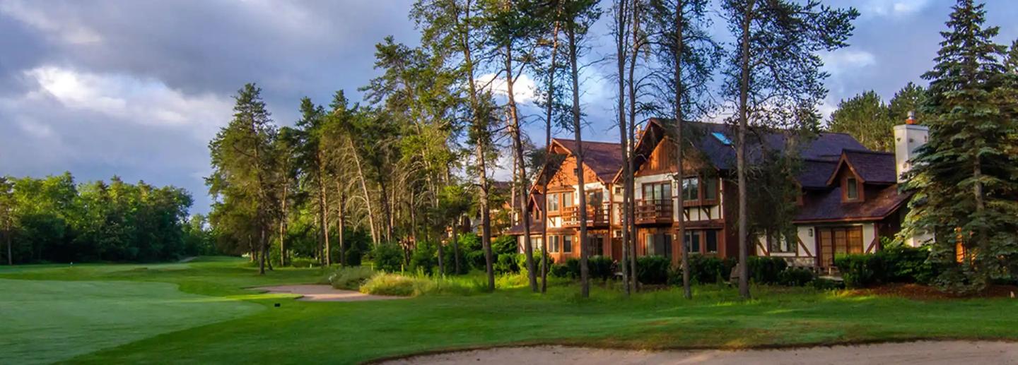 Large cabin surrounded by trees, next to a lush golf course under a cloudy sky.