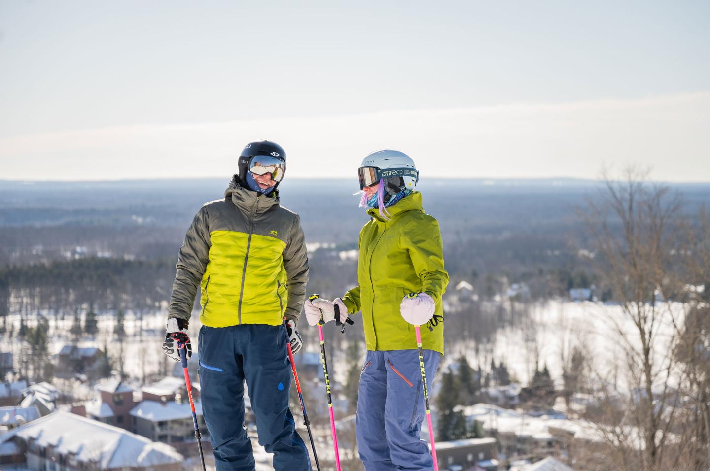 Two skiers in winter gear stand on a snowy hilltop with a forested valley below.