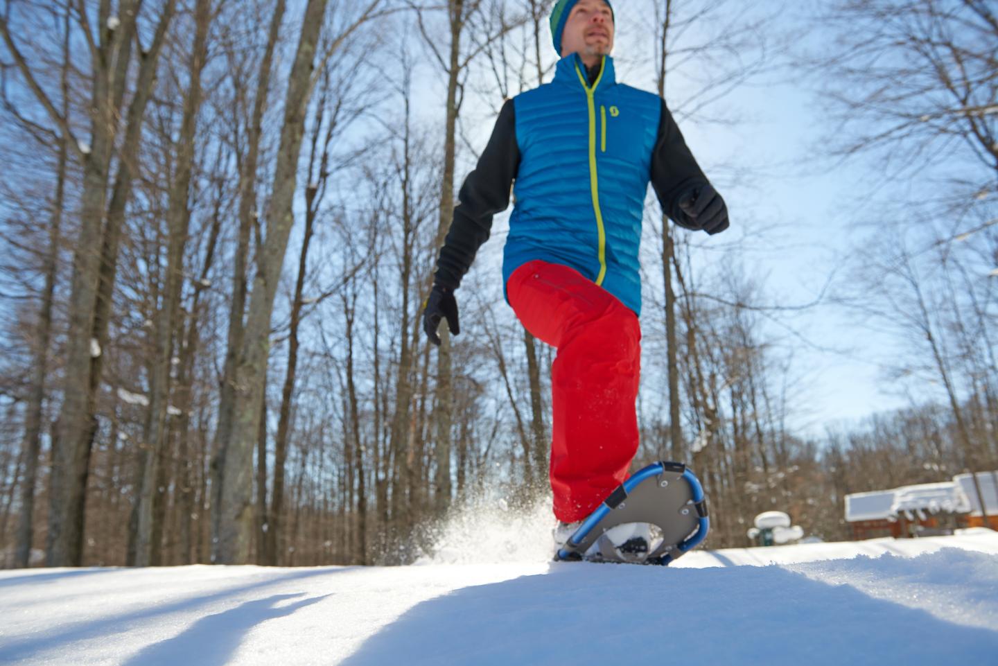 Man snowshoeing in winter forest, wearing blue vest and red pants.