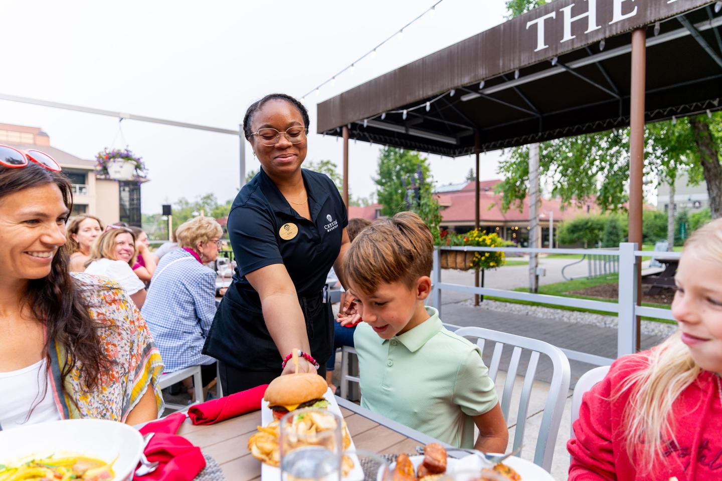 Server handing food to a smiling boy at an outdoor restaurant table.