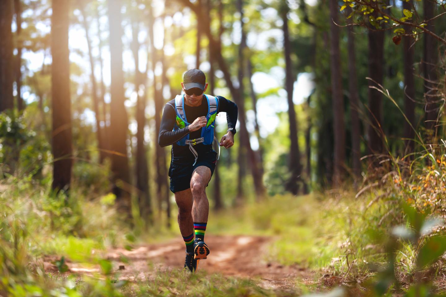 Trail runner in forest, wearing sunglasses and a hydration pack, surrounded by tall trees.