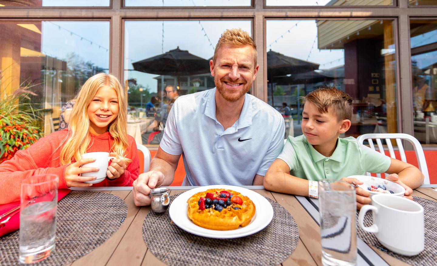 A man and two kids smile at a table with waffles and drinks outside.