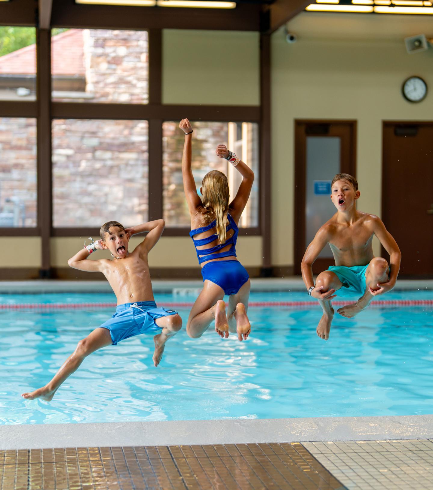 Three kids laughing and jumping into an indoor pool.