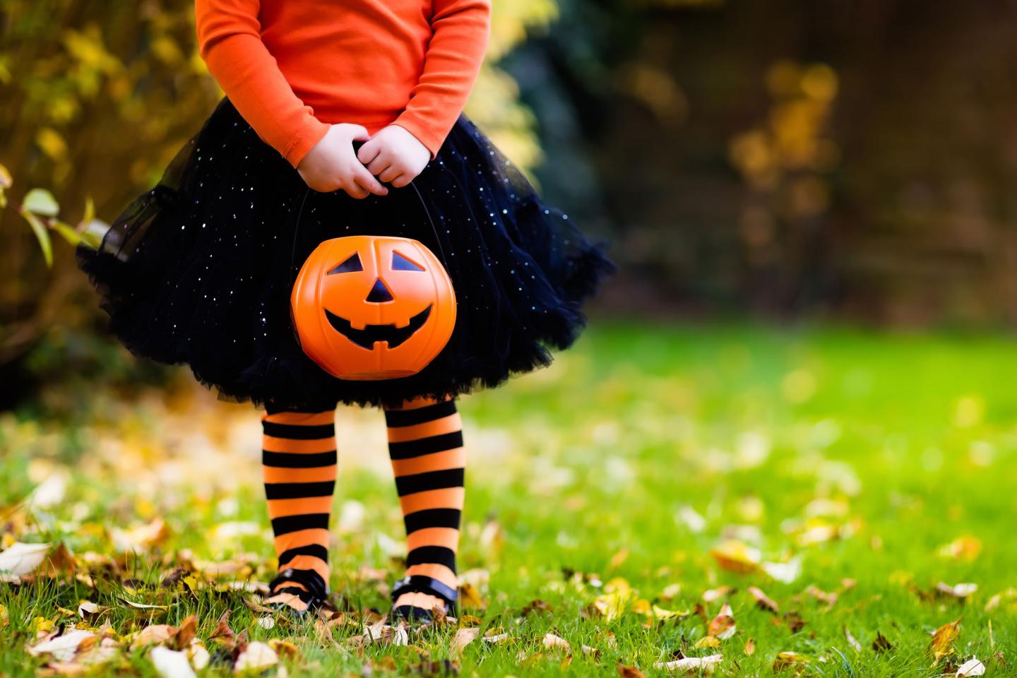 Child with a black skirt and striped tights holds a pumpkin basket on grass.