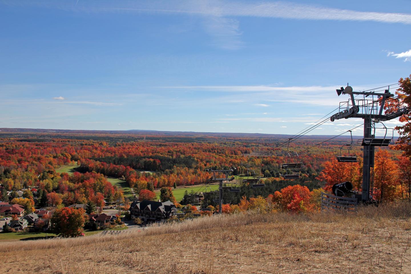 Autumn landscape with colorful trees, distant hills, and a ski lift under a clear blue sky.