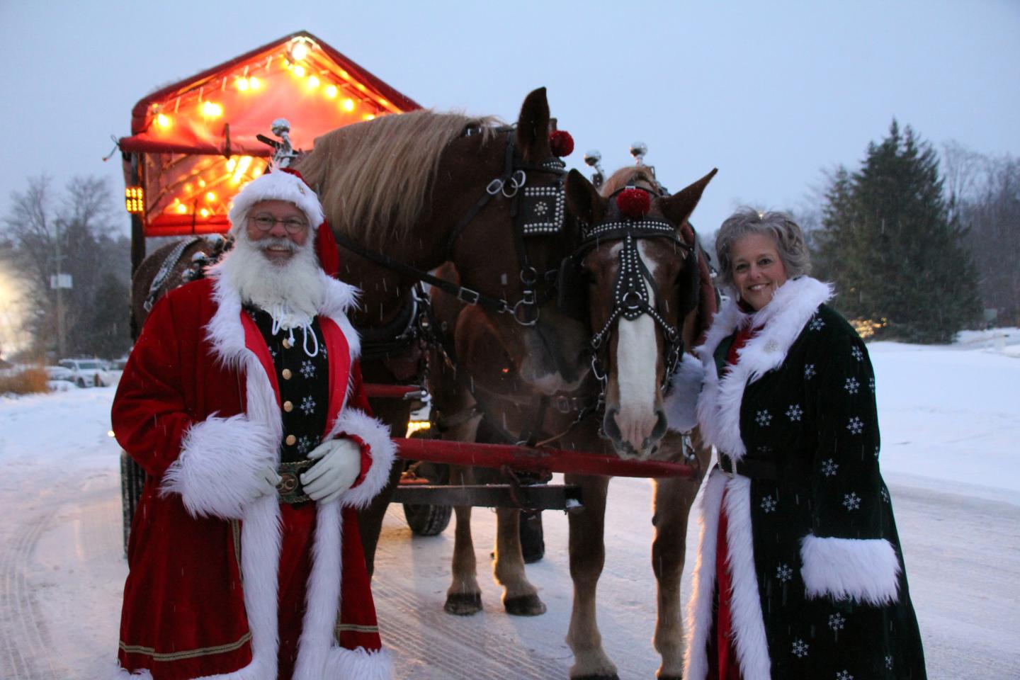 Santa and Mrs. Claus pose with horses and a lit sleigh in snowy setting.