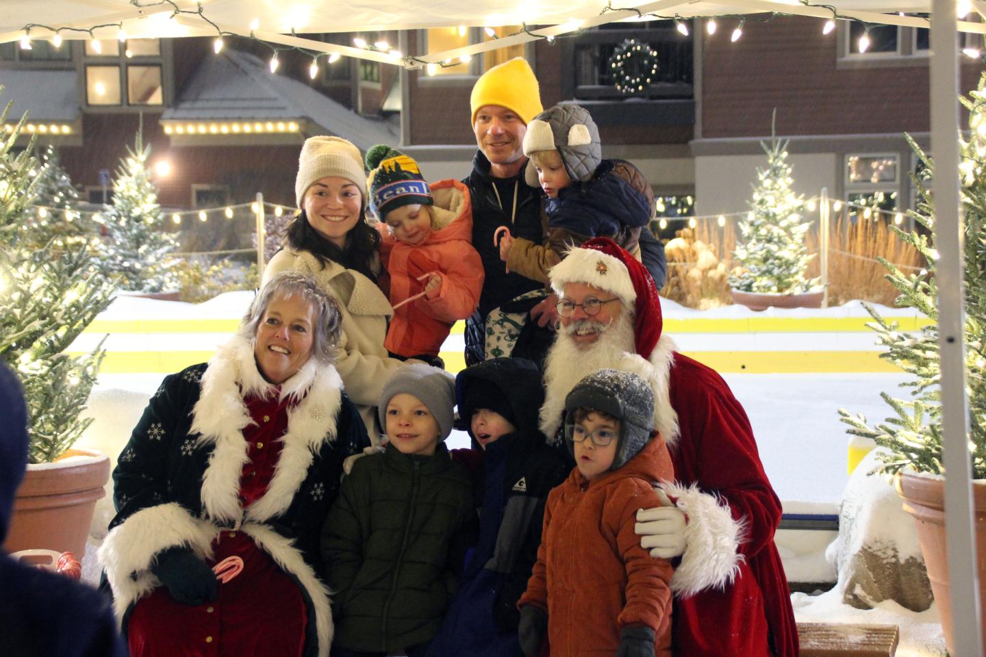 Santa and Mrs. Claus with a family outdoors, festive lights and trees in the background.