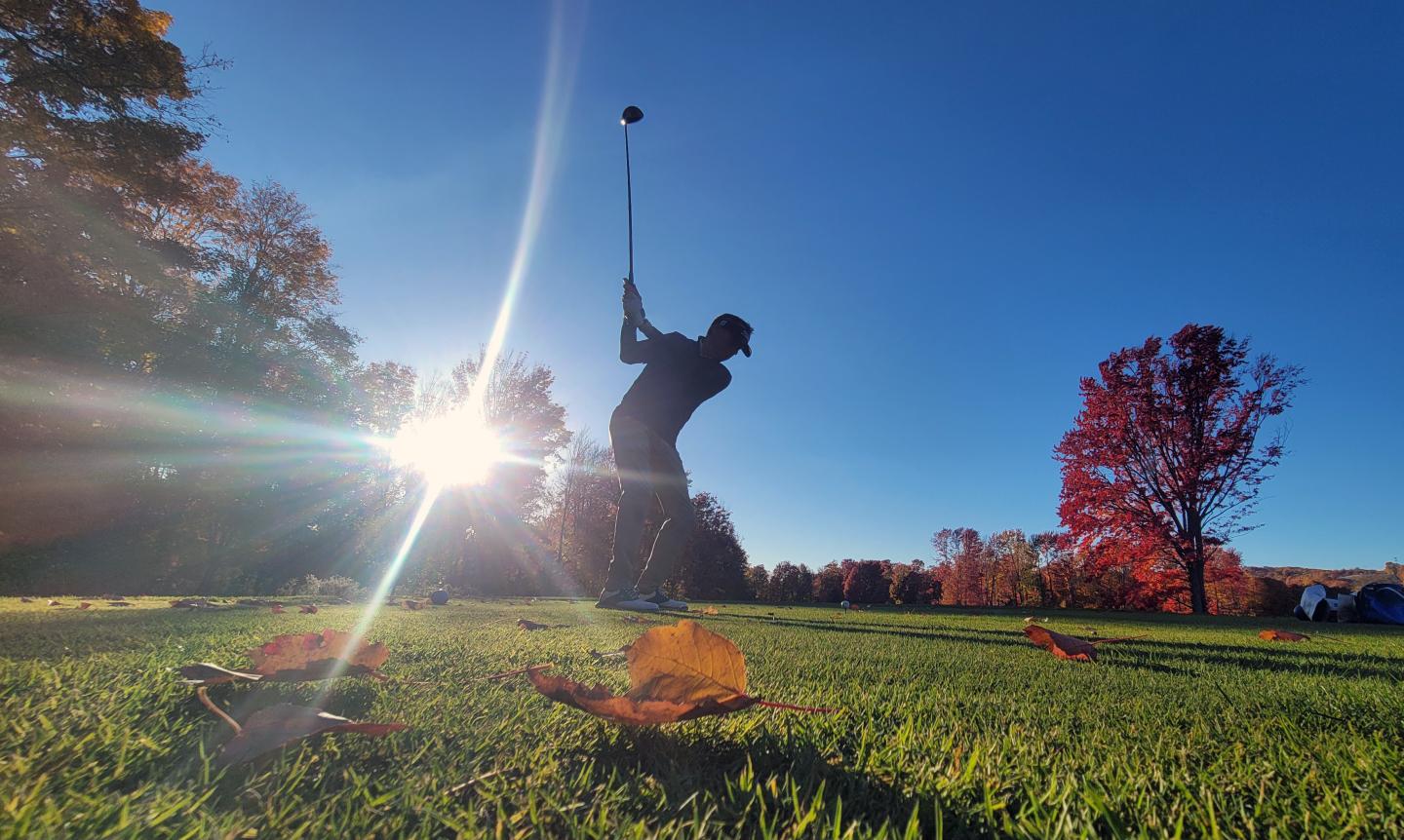 Golfer swinging under bright sun, autumn leaves on grass.