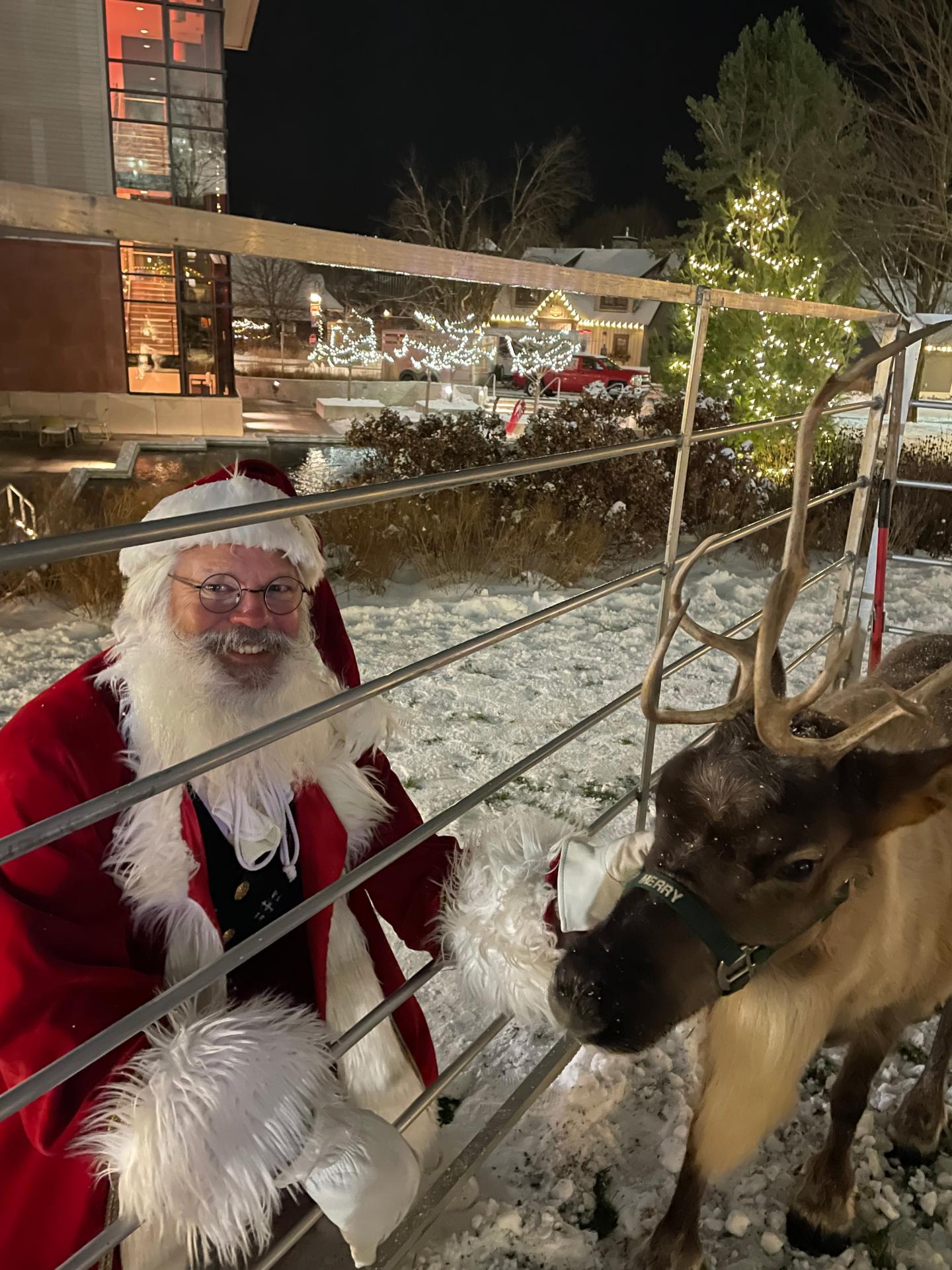 Santa with a reindeer in a snowy, festive setting at night.