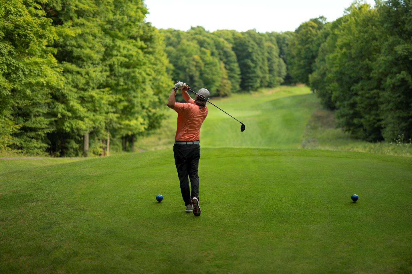 Golfer in orange shirt swings club on green course, surrounded by trees.