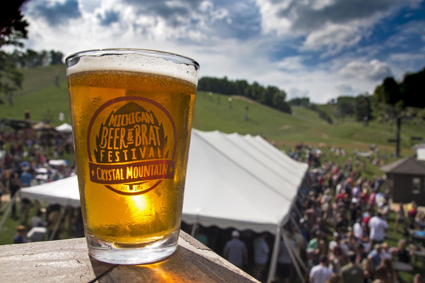 A beer glass with a logo, festival crowd in background under a blue sky.