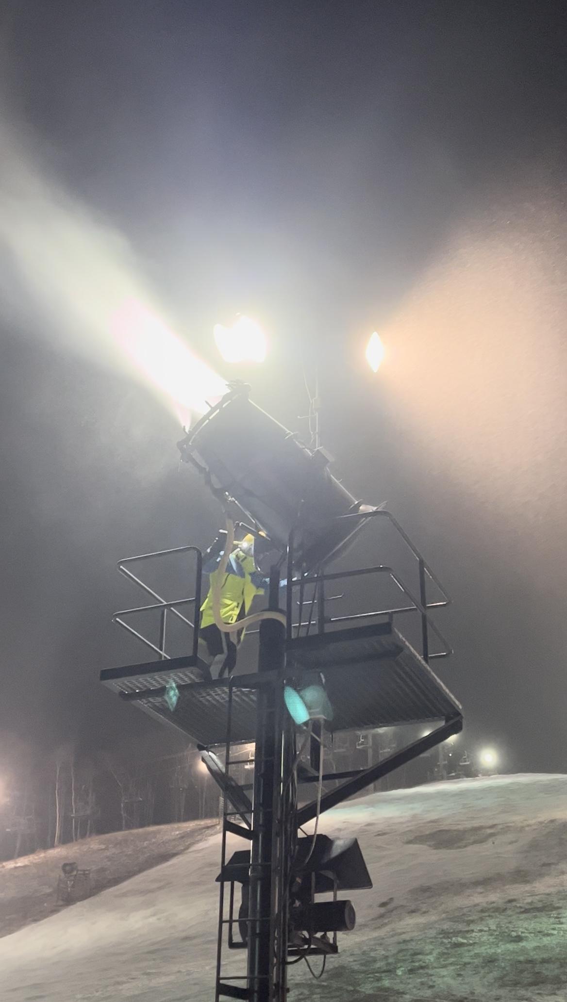 Snow machine operating on a ski slope at night, casting bright light and mist.