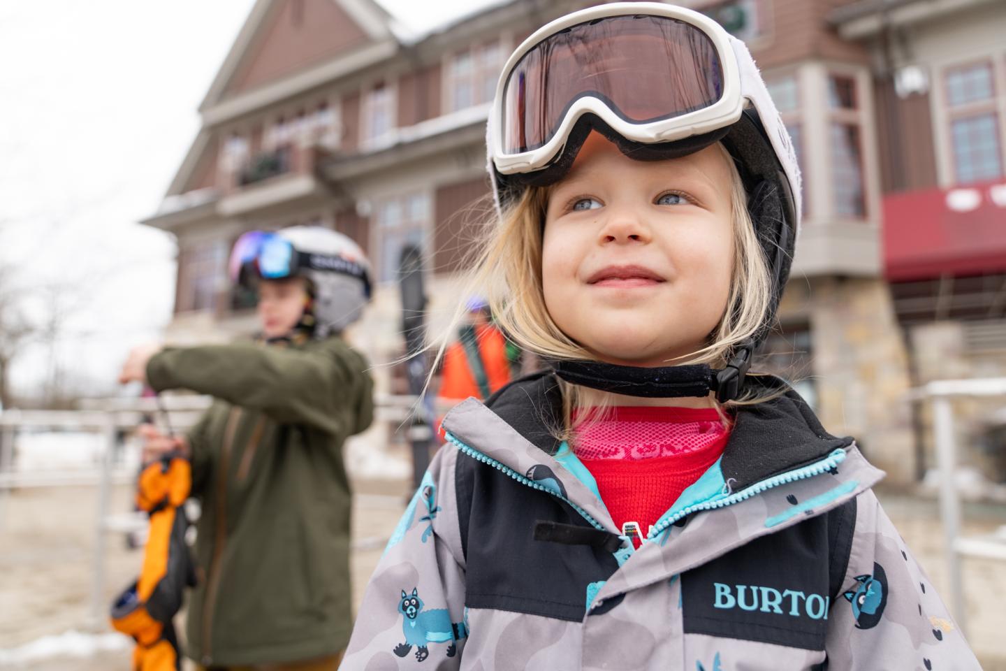 Young child in winter gear smiling, with skiers and lodge in the background.