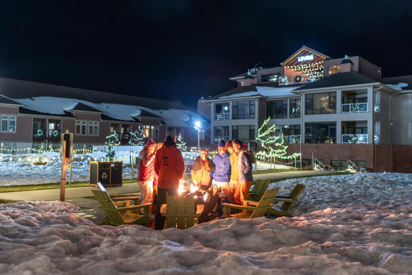 Group sitting around a fire pit at night, snowy resort background.