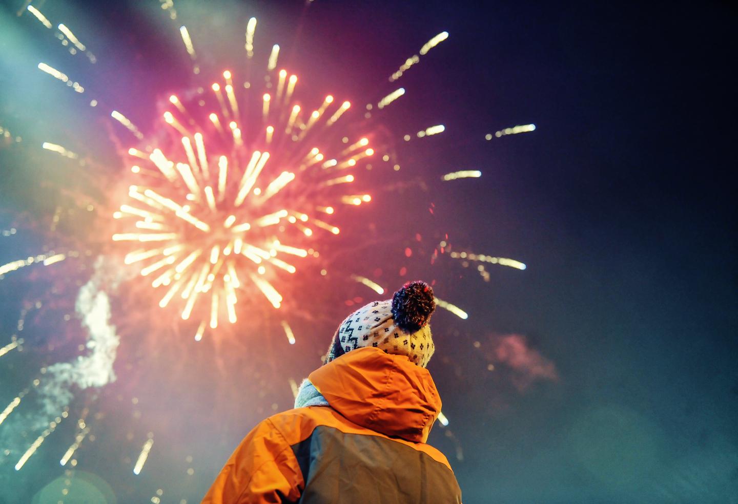 Child in winter clothing watches colorful fireworks in night sky.