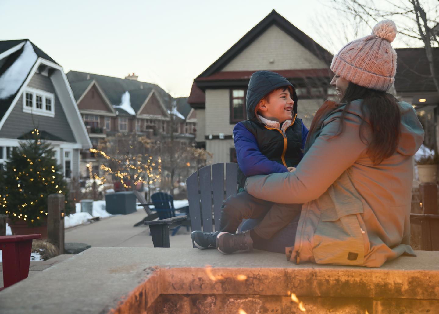 Mother and child smiling by a fire pit with holiday lights.