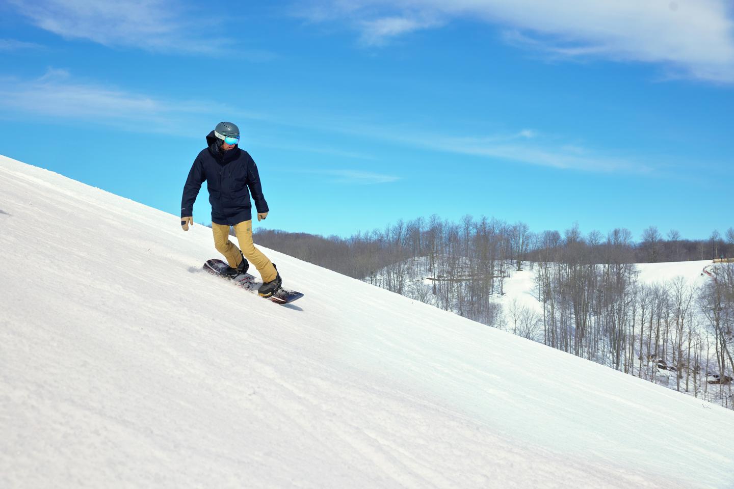 Snowboarder descending a snowy hill under a clear blue sky.