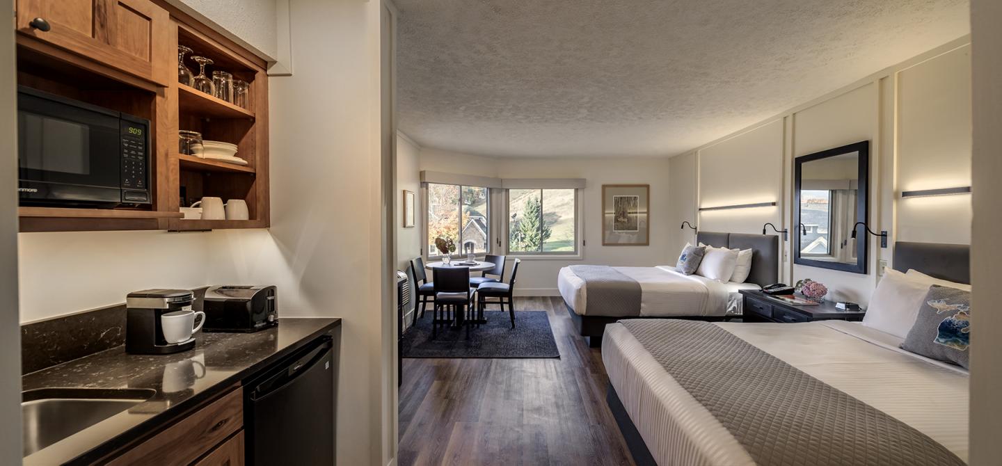 Hotel room with two beds, kitchenette, and a seating area by a window.