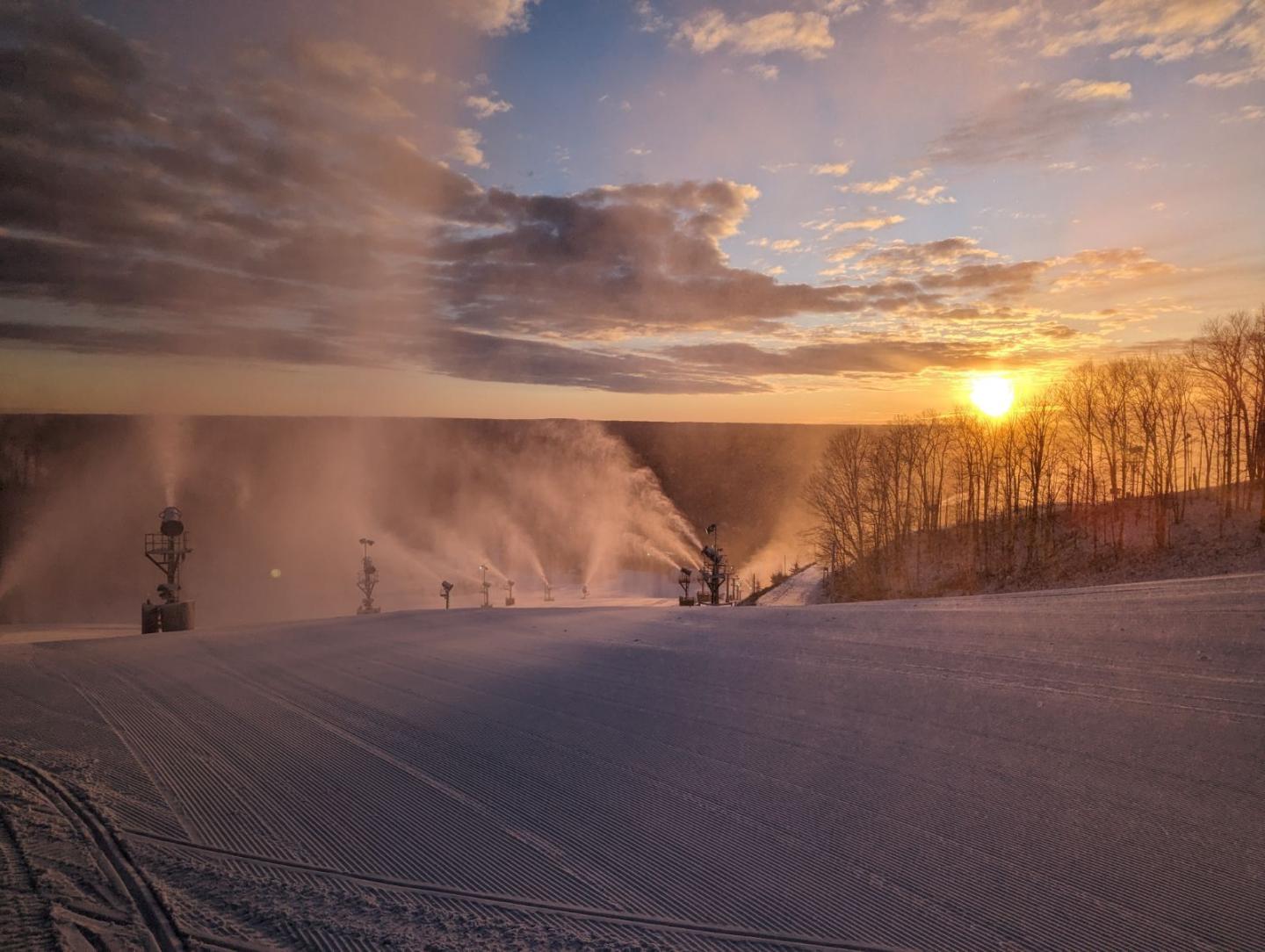 Sunrise over snowy landscape with snow machines and trees.