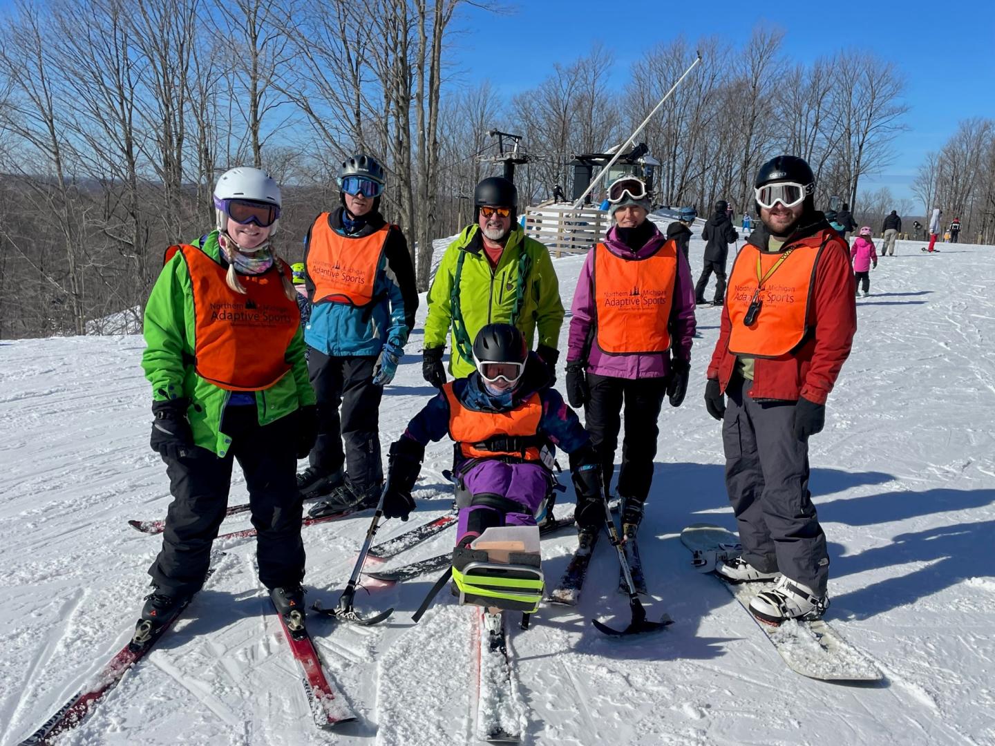 Group of NMAS skiers in colorful gear on a snowy slope, with trees and clear blue sky.