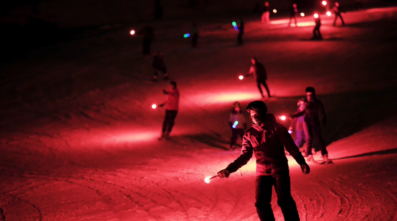 Skiers holding red lights on a snowy slope at night.