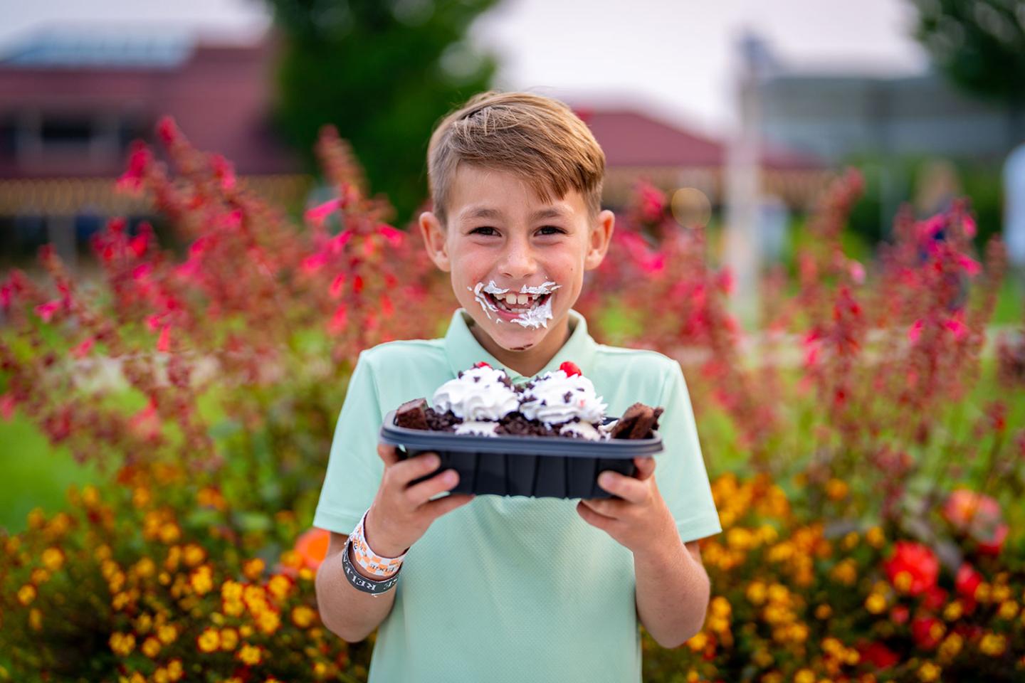 Boy smiling with whipped cream on his face, holding a dessert, colorful flowers behind.
