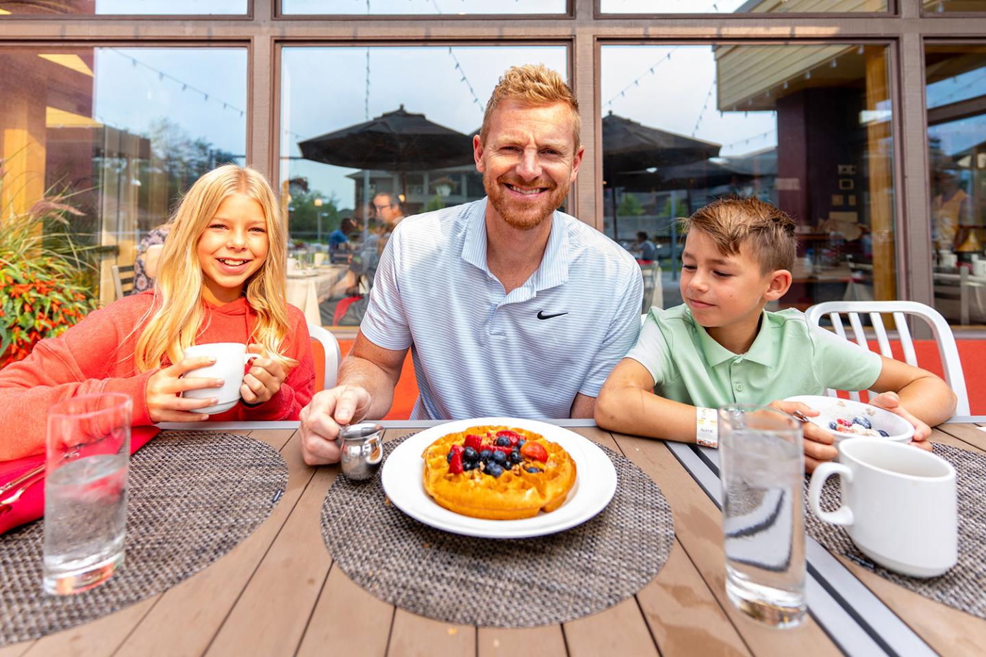 Man and two children enjoying breakfast outside, with a waffle and drinks on the table.