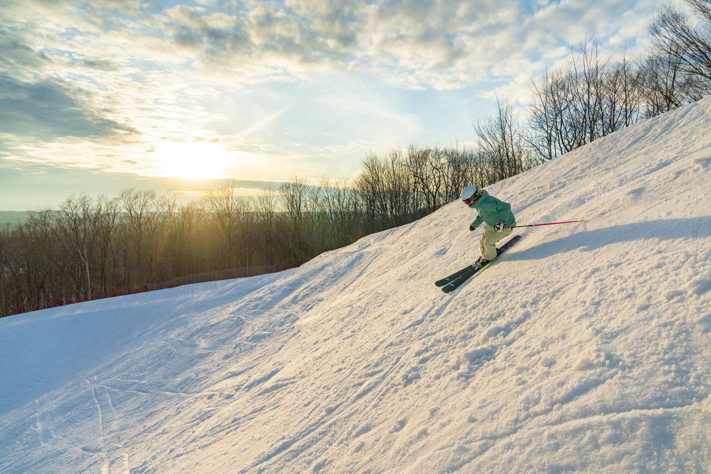Skier descending snowy hill at sunset with trees in the background.