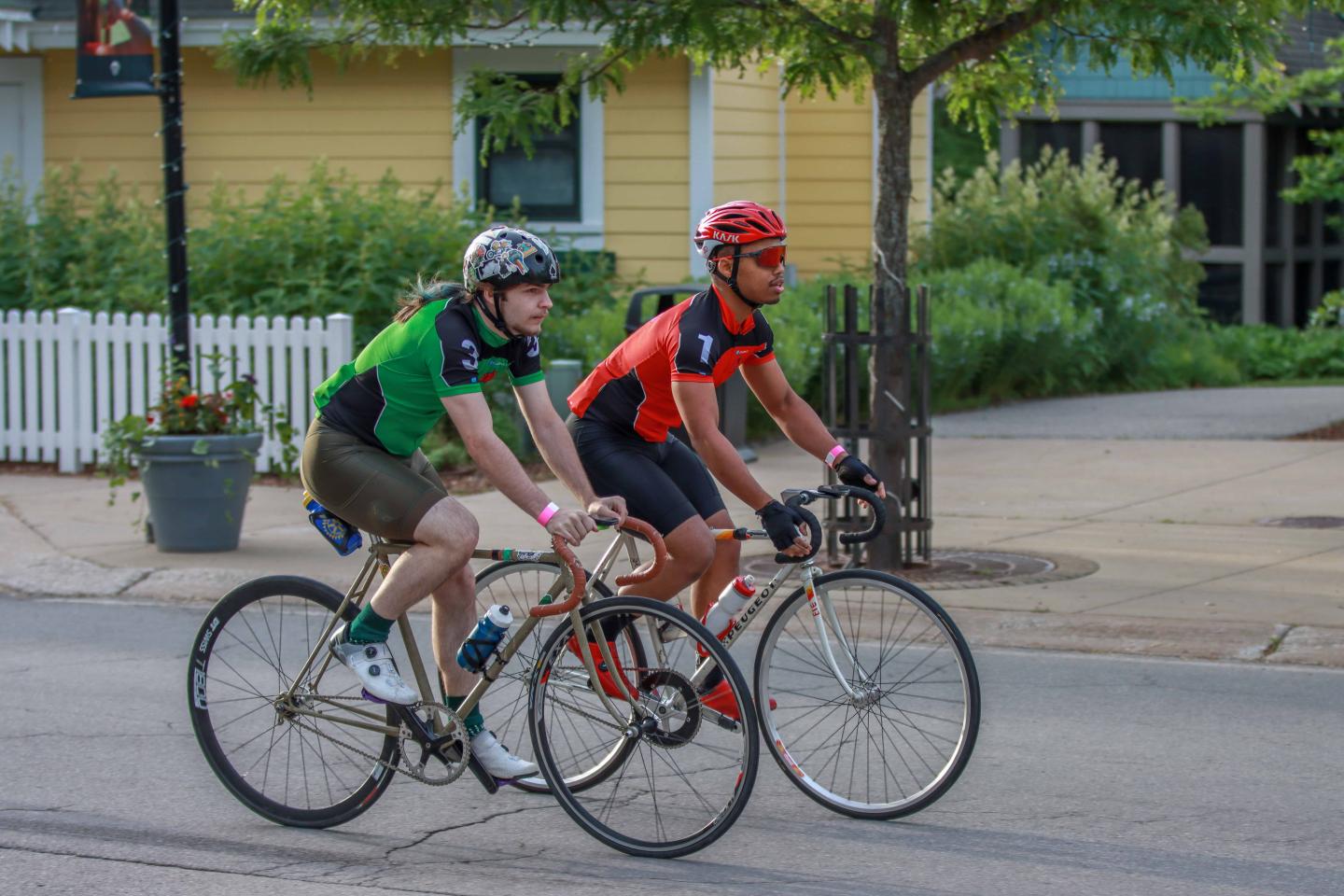 Two cyclists ride on a tree-lined street in summer gear, wearing helmets.