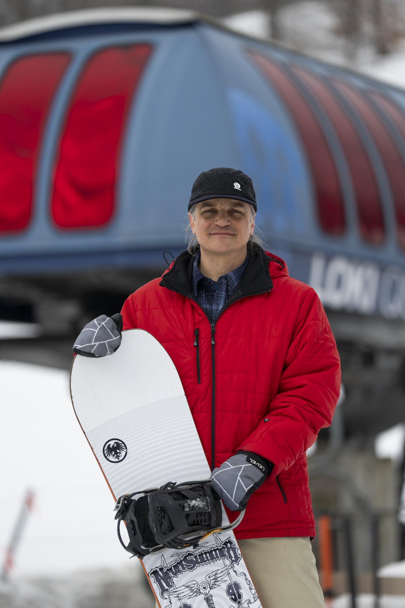 Man in red jacket and black cap holding a snowboard, standing by a ski lift.