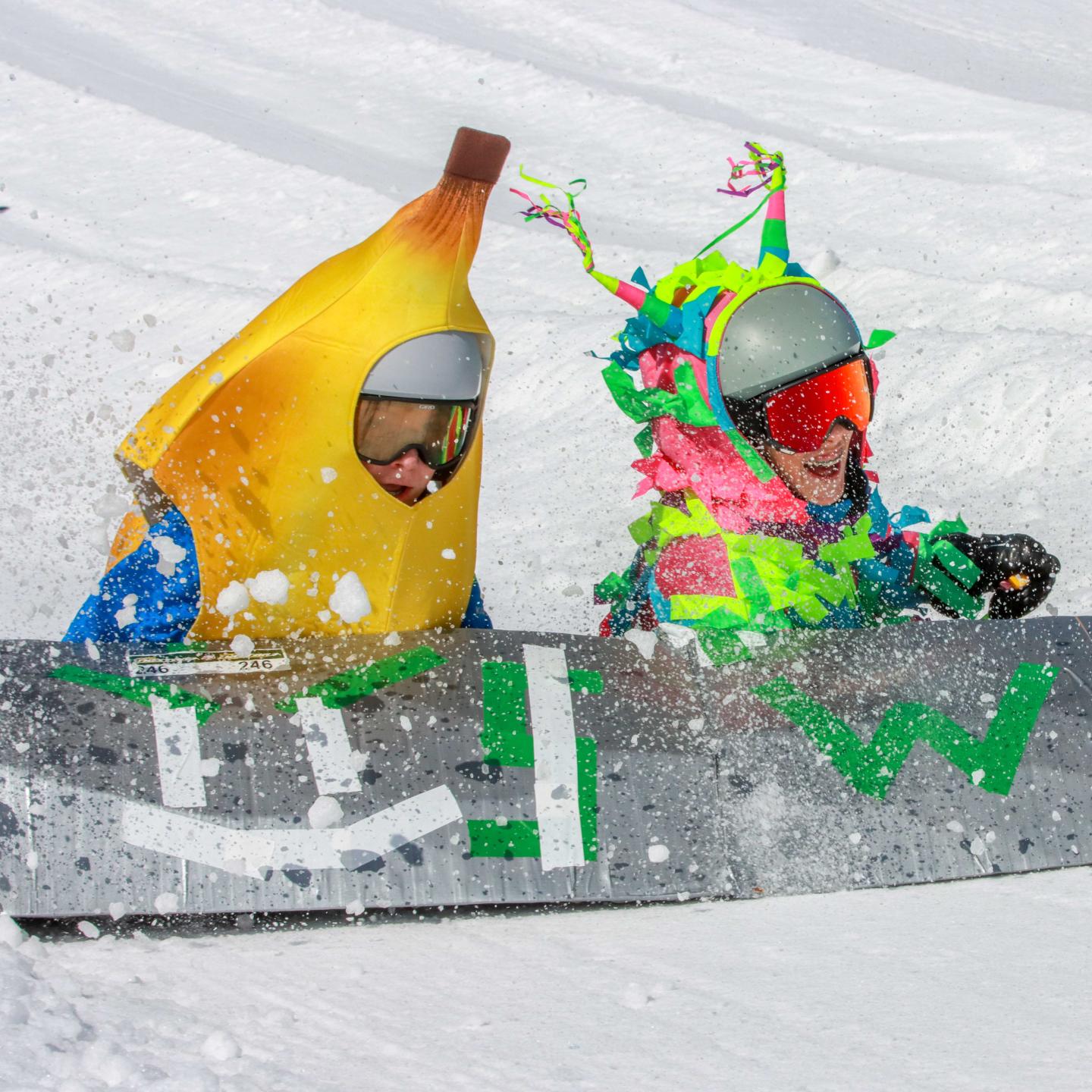 Two people in colorful costumes sledding in snow, one in a banana suit.